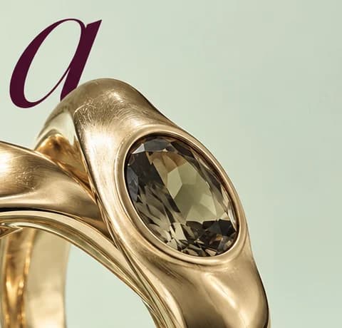 Close-up of a gold ring set with an oval smoky stone, its rim touching a plum italic letter a on pale green