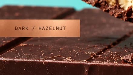 Copper foil strip reading DARK / HAZELNUT in monospaced capitals over a dark chocolate bar.