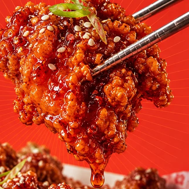 Glazed Korean fried chicken held by steel chopsticks, with sesame seeds, spring onion and a hanging drop of sauce.