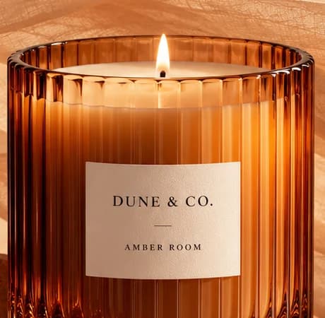 A lit candle in ribbed amber glass with a cream label reading 'DUNE & CO.' and 'AMBER ROOM', sheer gauze behind