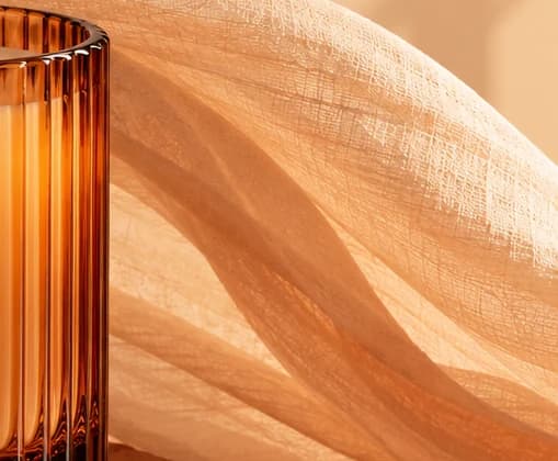 Side of the ribbed amber candle glass glowing in low sun, with sheer open-weave gauze curving behind it
