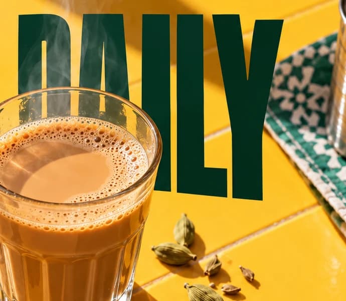 Karak glass in front of the green word DAILY, steam rising, with cardamom pods and a patterned cloth beside it.