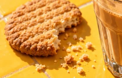 Round biscuit with a bite taken out, crumbs scattered on mustard tiles beside the base of the karak glass.