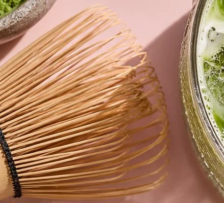 Bamboo matcha whisk on a blush pink tray, with a bowl of matcha powder and the glass at the edges.