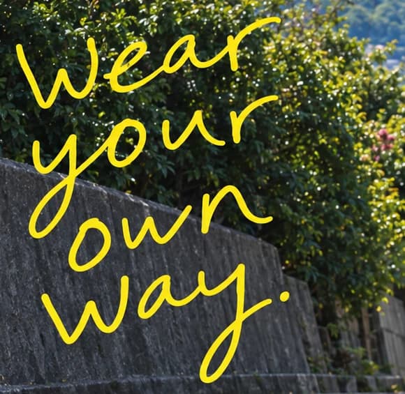 Soiu design close-up: Wear your own way. handwritten in yellow marker over a stone wall and green hedge