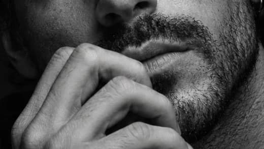 Close-up of a man's hand resting at his bearded chin, in high-contrast black-and-white light.